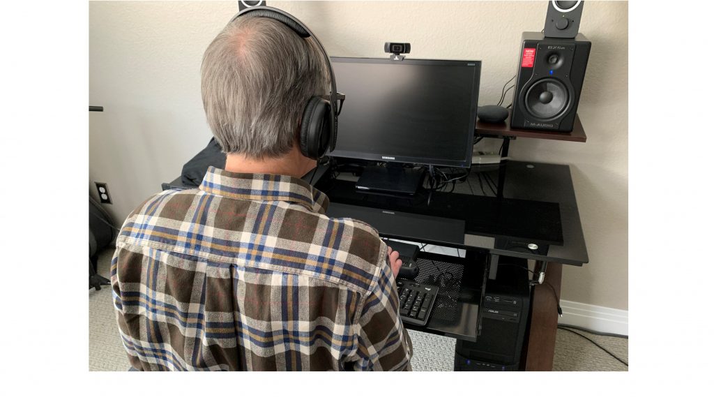 Blind man working at a computer from behind. Monitor is off; headphones are on; hands are on braille display