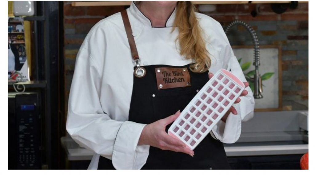 woman wearing a "the blind kitchen" apron holding an icecube tray to demonstrate using it to pre-measure ingredients
