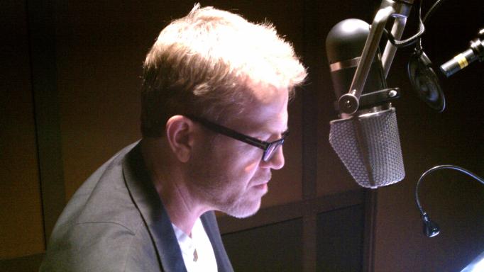 Scott Brick as seen from the side reading a book into a large microphone. The recording studio is fairly dark, but he has a bright light shining on the text he is reading from.