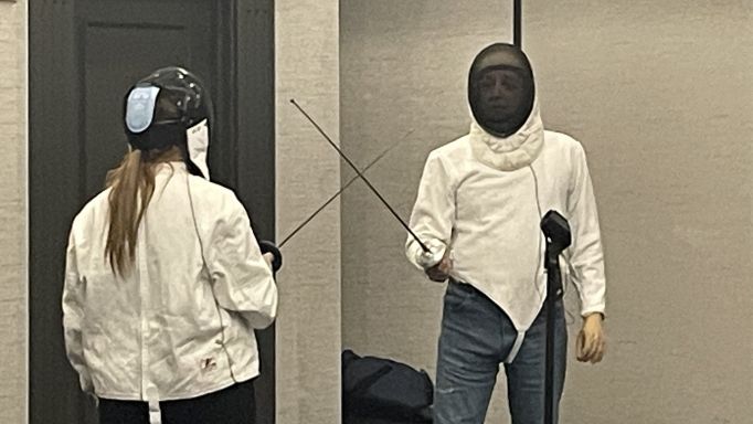 A photo of two people fencing. They are facing each other, each wearing a protective jacket and head shield. Each is holding a sword. The swords are pointed slightly upwards and are crossed in preparation for starting a round.