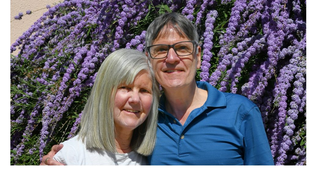 Nancy and Peter Torpey standing in front of a large butterfly bush covered in purple flowers on a sunny spring afternoon