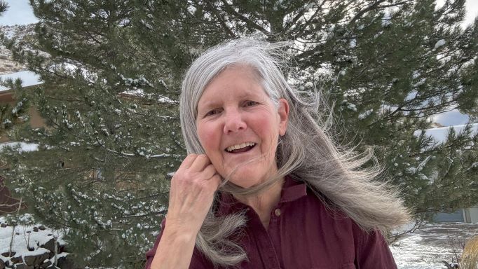 close-up photo of host Nancy Goodman Torpey with her long silver hair blowing in the breeze in front of a large pine tree on a snowy day