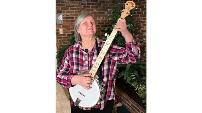 Sheri Wells-Jensen playing a banjo and wearing a plaid shirt in front of a brick wall