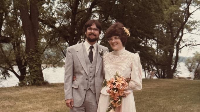 Pete and Nancy Torpey's wedding photo. They are smiling broadly with their arms around each other's waists and standing on a grassy lawn with large lush trees and Irondequoit Bay behind them. He is wearing a 3-piece gray suit with a white shirt, a dark gray tie and a white boutonniere. She is wearing an off-white chiffon and lace dress and holding a bouquet of peach colored lilies and gladioli with a single lily blossom in her hair.
