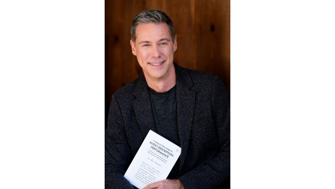 photo of Roy Samuelson holding his book 'Audio Description Performance'. He is wearing a dark sport jacket with a dark crew-neck shirt and is in front of a dark wood wall.