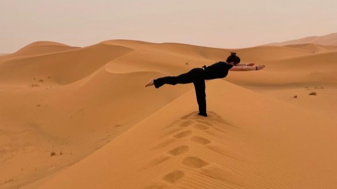 photo of Aly Slaughter in Warrior 3 position in the Sahara desert. She is standing on her left foot which is buried in sand up to the ankle. Her right leg and both arms are horizontal, forming a capital T with her body. She is wearing all black and is very visible against the tan sand dunes.