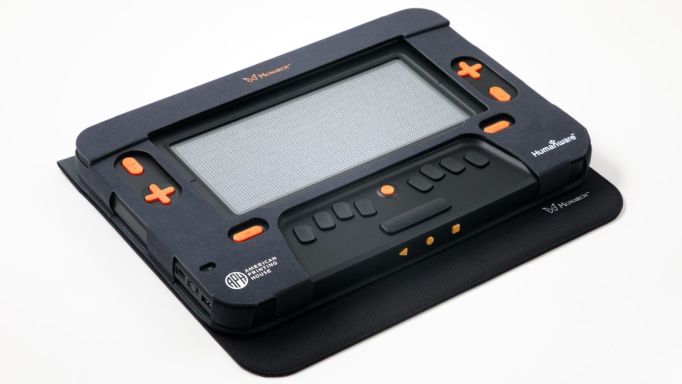 Photo of a Monarch 2D display. It is black, about the size of a typical book, and has large raised pale orange buttons for controls. It also has ports for connectivity on the edges.