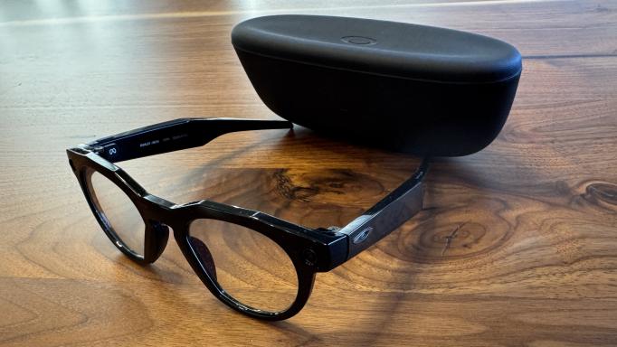 Photo of black framed Oakley Meta Glasses with the case which is matte black. Both are sitting on a dark wood table.